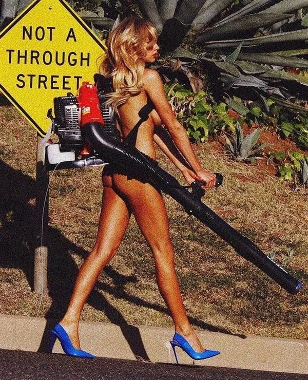 A woman in high heels and revealing clothing uses a leaf blower while walking past a NOT A THROUGH STREET sign on a sunny street with greenery in the background.