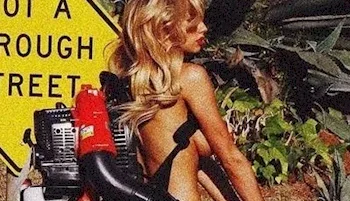 A woman with long blonde hair, wearing only a backpack leaf blower, stands outside near a yellow road sign and green foliage.