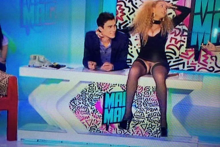 Two people sit at a colorful TV show desk. The woman in a black outfit and stockings sits on the desk, while the man in a suit sits beside her. The background displays bold, graphic art with MA MA text.