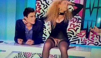 Two people sit at a colorful TV studio desk; the woman in a revealing outfit adjusts her hair, while the man beside her smiles, facing forward. Bold, patterned wall art is visible in the background.