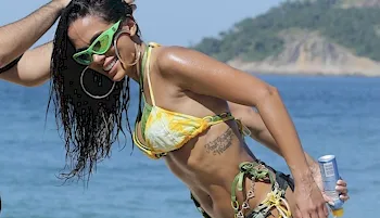 Anitta in a bikini and bending over at the beach with a huge smile on her face