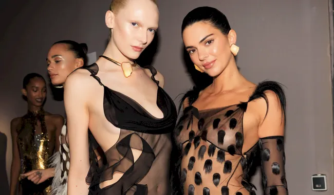 Kendall Jenner braless in a sheer dress posing next to another model behind the scenes at a fashion show