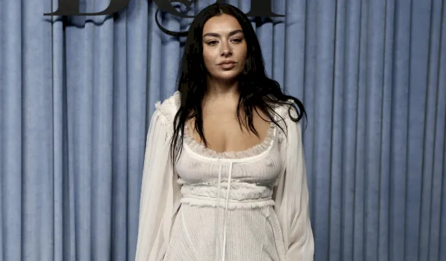Charli XCX in a white see through dress posing in front of blue curtains