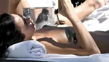 Dua Lipa in a bikini and laying on her back in a sunchair reading