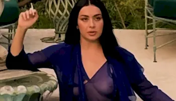 Charli XCX holding a cigarette while posing in a sheer and purple dress