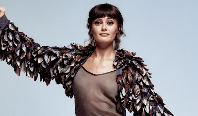 Close up of Ella Purnell posing with one arm raised in a sheer top and with feathery sleeves