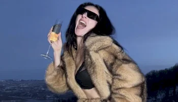 Charli XCX holding a champagne glass and smiling big while wearing a big fur coat