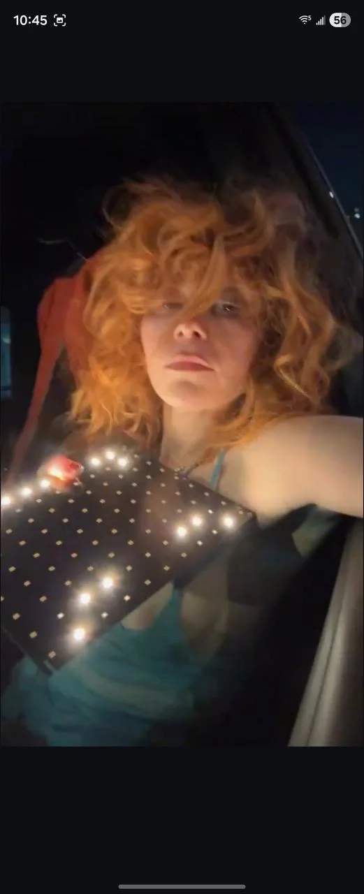 A person with curly red hair is sitting in a dimly lit space, wearing a teal top and holding a rectangular object with small lights and a red button. The scene appears to be taken at night.