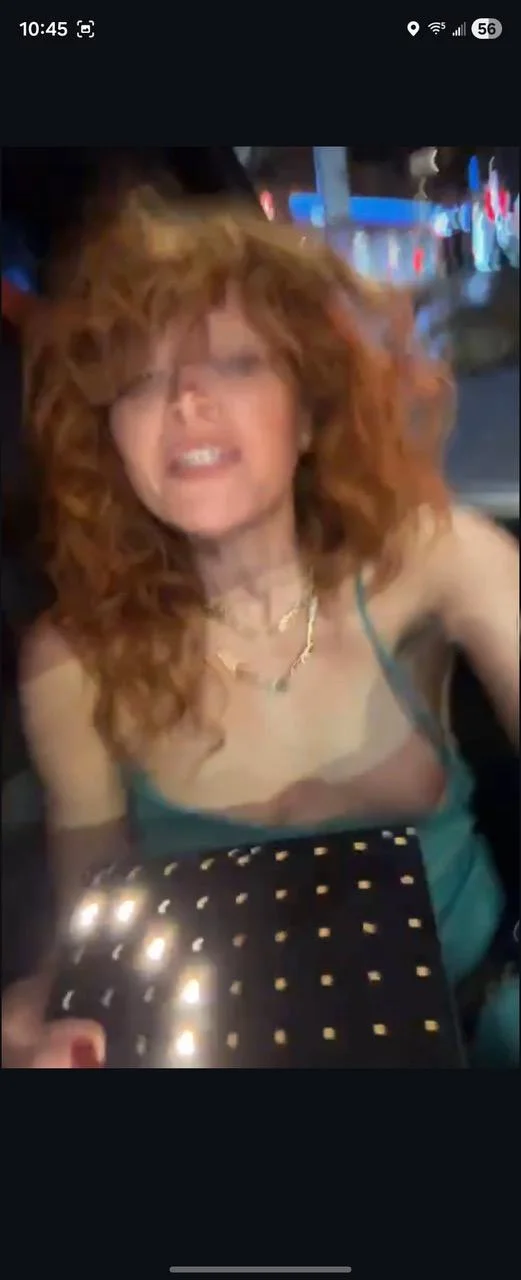 A person with long, curly red hair wearing a turquoise top and layered necklaces holds a black box with lit-up dots. The image is taken at night and appears slightly blurred.
