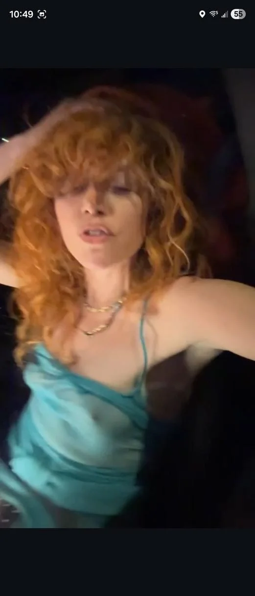 A woman with long curly red hair wearing a turquoise spaghetti strap top poses with one hand in her hair, looking slightly away from the camera. The background is dark and out of focus.