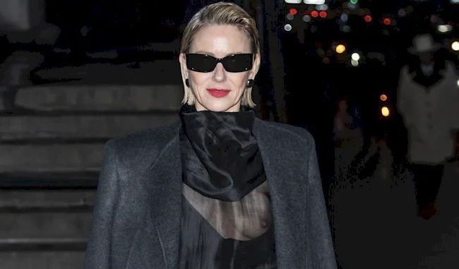 A woman with short, slicked-back blonde hair wears black sunglasses, a dark sheer top, a dark jacket, and bright red lipstick while standing outdoors at night with city lights in the background.