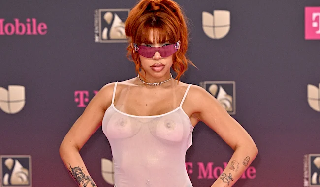 A person with reddish hair in a ponytail poses on a pink carpet, wearing large purple sunglasses, a sheer white dress, and displaying visible tattoos on their arms. Pink and gray logo backgrounds are behind them.