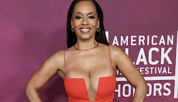 A woman in a red dress poses, smiling, with hands on hips in front of a purple backdrop that reads American Black Film Festival Honors.