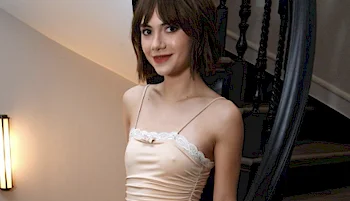A young woman with short brown hair and bangs is standing on a staircase, wearing a light-colored satin dress with thin straps and lace trim, smiling softly at the camera.