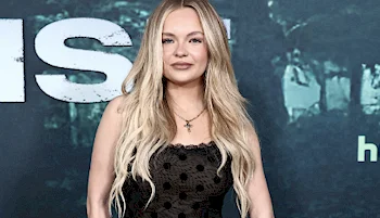 A woman with long, wavy blonde hair wearing a black, sleeveless, polka-dot dress stands in front of a dark blue and green backdrop with bold white lettering.