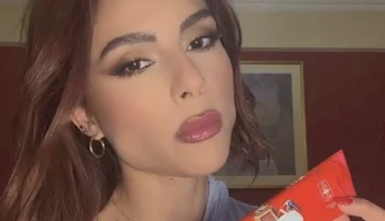 Close up of woman with wavy brown hair and bold makeup holds a red chocolate bar package, looking into the camera with a neutral expression. A painting hangs on the wall behind her.