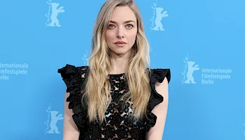 A woman with long blonde hair wearing a black lace dress stands in front of a blue backdrop with white logos for the Berlin International Film Festival.