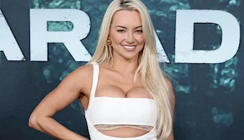 A woman with long blonde hair wearing a white cut-out dress stands in front of a dark backdrop with large, partially visible white letters. She is smiling and posing with one hand on her hip.