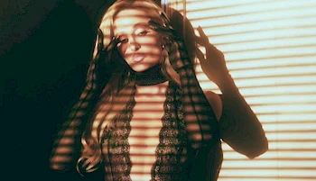 A woman in a lacy outfit stands in dramatic lighting, with shadows from window blinds casting striped patterns across her face and body. She has long blonde hair and gazes directly at the camera.