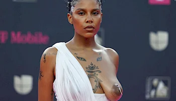 A woman with short hair and tattoos wears a white asymmetrical dress that exposes one side of her chest. She stands on a red carpet with a serious expression, and a blurred backdrop features logos and text.
