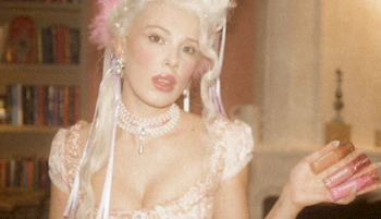 A woman in a Marie Antoinette-inspired costume with a powdered wig, pearl necklace, and pink dress stands indoors, holding what appears to be small bottles or tubes in her hand. The image has a soft, vintage look.