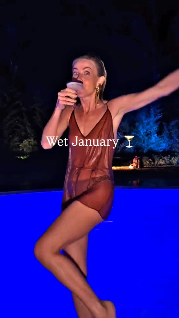 A woman in a sheer, wet dress stands by a glowing blue pool at night, holding a drink with arm more raised.