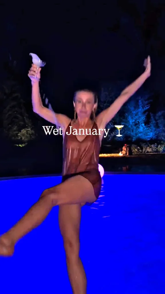 A woman in a sheer, wet outfit, holding a drink, is falling backwards into a glowing blue pool at night.