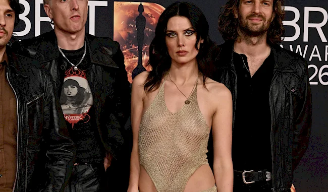 Three people posing on a red carpet at an event. The person in the center wears a revealing gold dress, while the two people beside them wear dark jackets. The background has event signage.