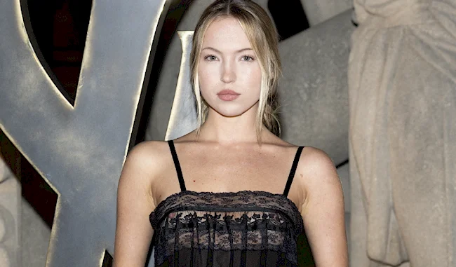 A woman with long blonde hair, wearing a black lace dress, stands in front of a backdrop featuring large stone sculptures and the letters Y and L.