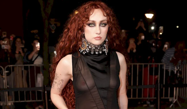 A girl with long, curly red hair wears a sheer black sleeveless dress and a large, ornate choker necklace, standing outdoors at night with people and cameras in the background.