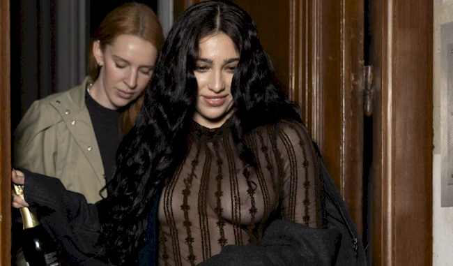 Two women exit a building; the woman in front has long black hair and wears a sheer black top with vertical stripes, holding a dark jacket. The woman behind her has long red hair and wears a beige jacket, holding a bottle.