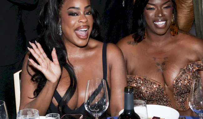 Two women dressed in elegant evening wear sit at a dinner table, smiling and waving, with glasses and a bottle visible in front of them.