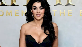 A woman with long, wavy black hair and gold earrings poses in a strapless black dress in front of a light backdrop with gold lettering. She is smiling at the camera.