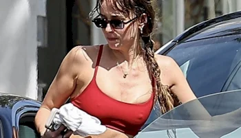 A woman wearing sunglasses and a red sports bra stands next to a car, holding a white piece of clothing. Her hair is tied back, and she appears to be outdoors in daylight.
