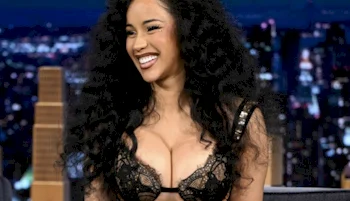 A woman with long, curly black hair is smiling while wearing a black lace top. She is sitting in a studio setting with city lights blurred in the background.