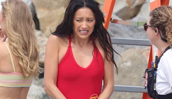 A woman in a red swimsuit appears cold or uncomfortable, grimacing and hunching her shoulders, while standing outdoors near a ladder and talking to two other women. Rocks are visible in the background.