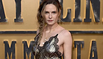 A woman with wavy hair styled in a loose side braid poses in a lace-trimmed metallic dress in front of a backdrop with large, bold text at a formal event.