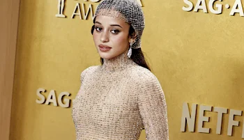 A woman poses on a red carpet wearing a sparkling nude-colored gown and a mesh headpiece, standing in front of a gold backdrop with SAG AWARDS and NETFLIX logos.