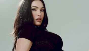 A woman with long dark hair and light eyes poses against a plain background, wearing a black top and looking confidently at the camera.
