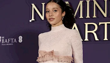 A woman wearing a high-neck, long-sleeved, light pink lace dress with a ruffle detail stands in front of a purple backdrop with large gold text and BAFTA logos.