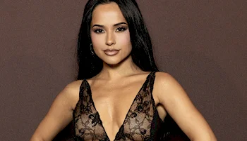 A woman with long dark hair poses confidently against a brown background, wearing a sleeveless, deep V-neck, sheer black lace top with floral patterns.