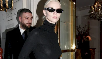 A woman with blond hair tied back, wearing black sunglasses and a black turtleneck, stands in front of a glass door. A man in a suit is visible behind her indoors.