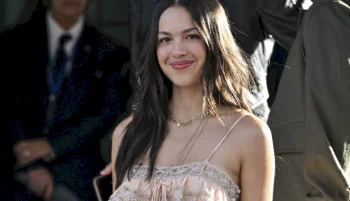 A young woman with long dark hair wearing a light pink spaghetti strap top stands outdoors, smiling at the camera. There are people and shadows visible in the blurred background.