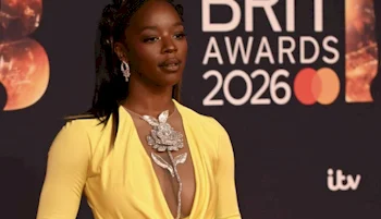A woman in a yellow dress with a silver floral necklace poses on the red carpet at the BRIT Awards 2026, with the event logo visible in the background.