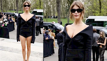 A woman wearing a black, off-the-shoulder bodysuit and dark sunglasses stands outdoors at a public event, with people seated and trees visible in the background.