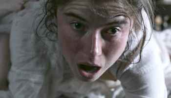 A woman with messy hair and a flushed face looks down with wide eyes and an open mouth, appearing shocked or distressed, while wearing a white shirt.