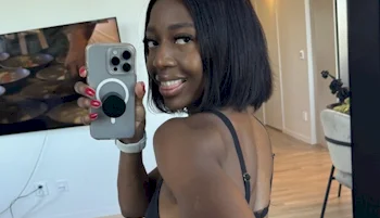 A black woman with a bob haircut smiles at the camera while taking a mirror selfie with her phone in a modern, brightly lit room. She is wearing a black top and has red nail polish.