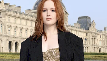 A woman with long reddish hair wearing a black blazer and a gold lace top stands in front of an ornate historic building with a domed roof, likely a famous museum.