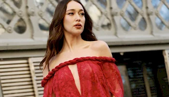 A woman with long brown hair wears an off-the-shoulder red lace dress with a braided neckline, standing outdoors in front of an ornate metal structure.