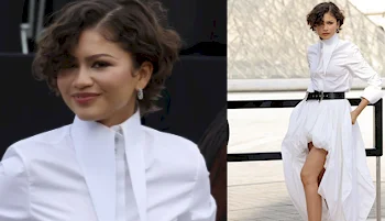 A woman with short, curly hair wears a high-collared, long-sleeved white shirt dress with a black belt and asymmetrical hem, posing confidently outdoors.